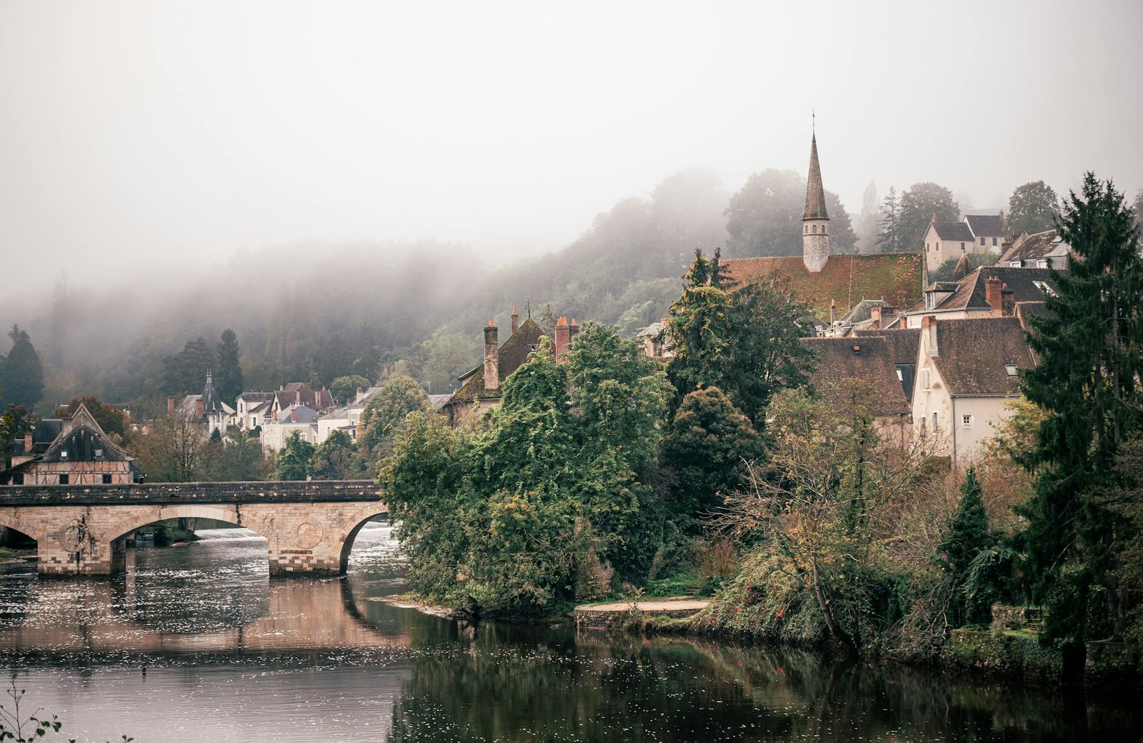 Charming misty morning view of a French village with a historic stone bridge over a river.