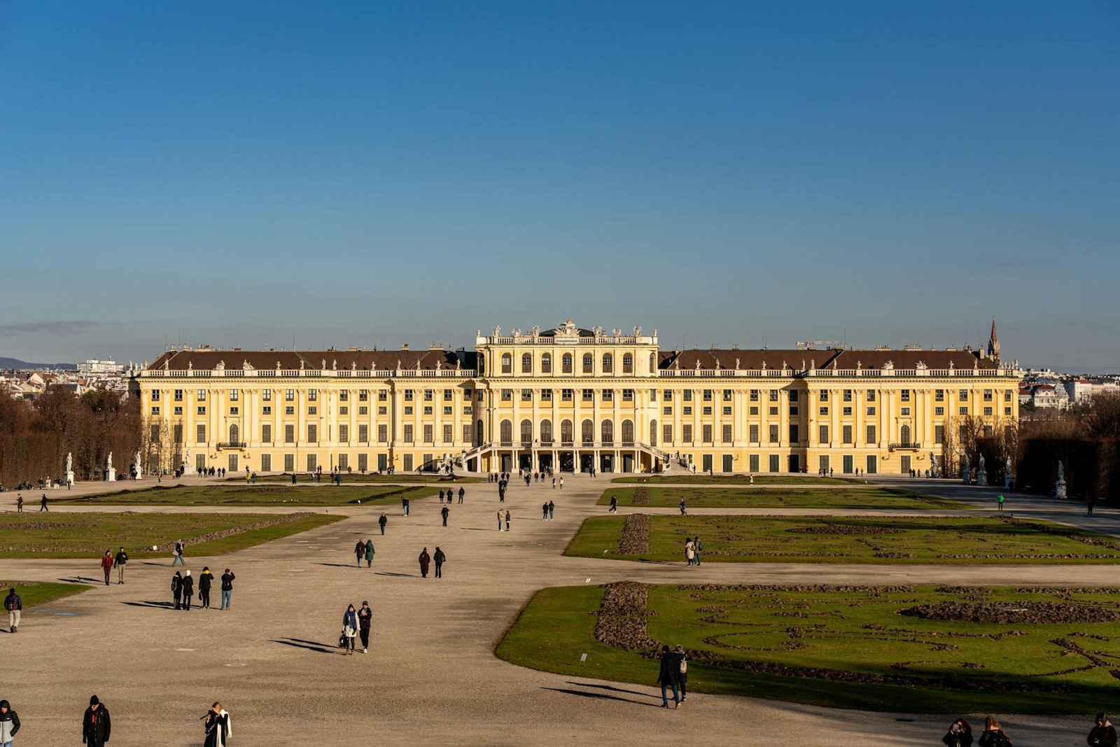 A frontal view of Schönbrunn Palace in Vienna, Austria, with people strolling the gardens.