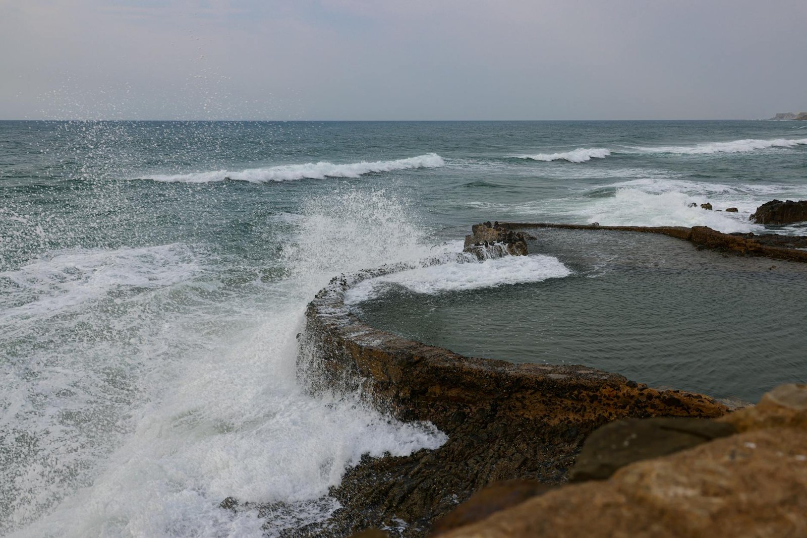 Dynamic waves crash against a natural rock pool on a cloudy day at the seaside.